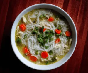 A bowl of Chicken Rice Noodle Soup topped with fresh herbs, sliced chicken, and lime wedges, served hot with colorful vegetables and rice noodles.