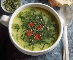 A steaming bowl of Caldo Verde soup featuring shredded kale, chorizo slices, and potatoes in a clear broth, served with crusty bread on the side.